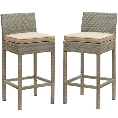 Pair of gray woven wicker outdoor bar stools with beige cushions, square legs and built-in footrests.