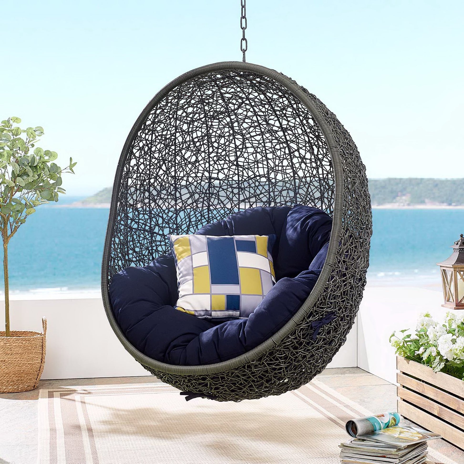 Dark gray fabric swing outdoor patio lounge chair without stand with navy cushions and a geometric blue-yellow pillow on a seaside patio.
