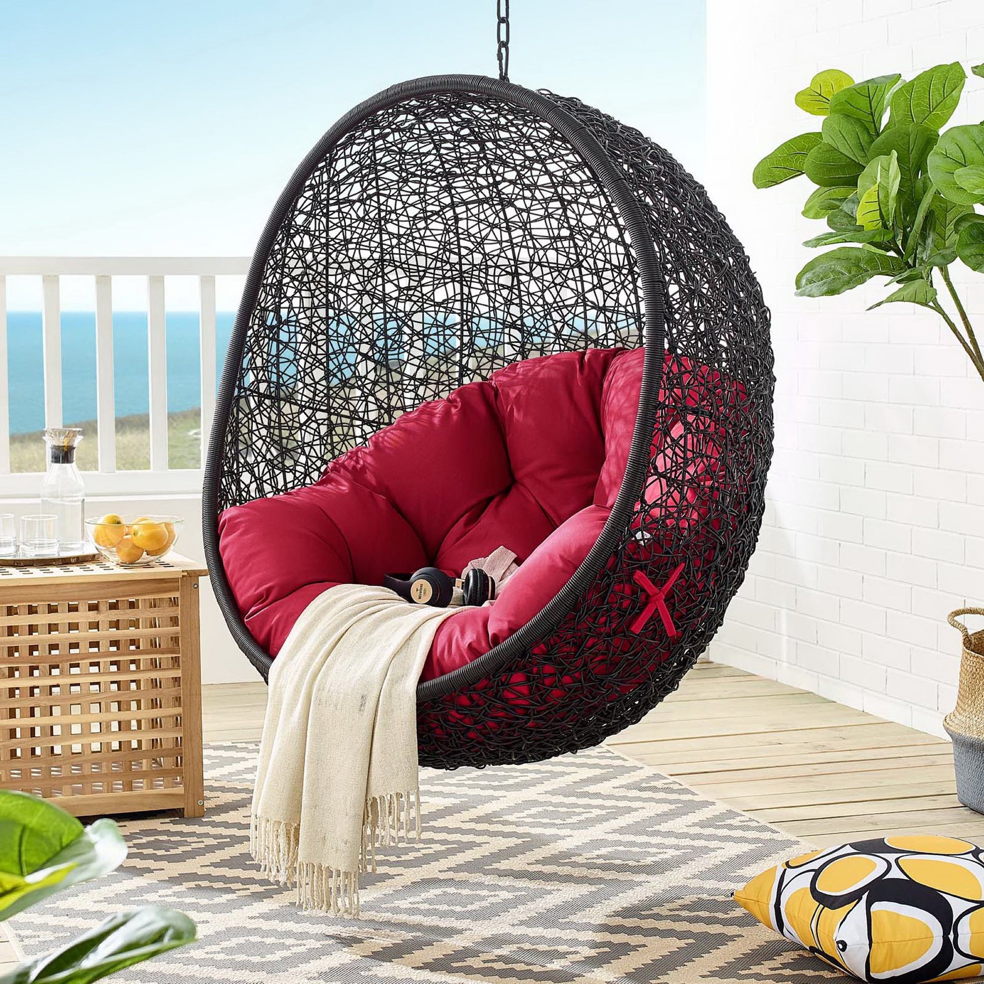 Black woven hanging egg chair with red cushions on a seaside balcony, cream throw draped inside
