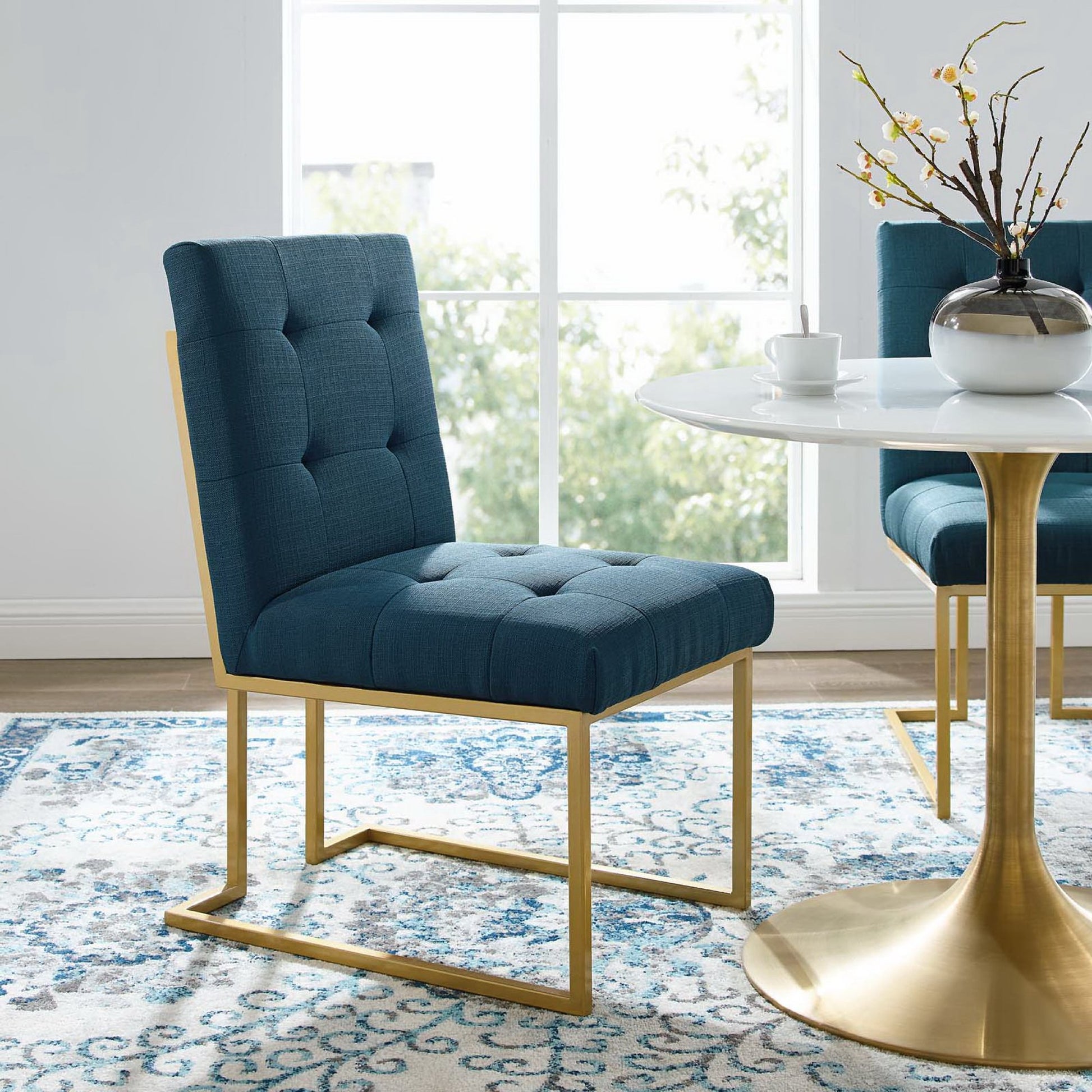 Azure tufted armless chair with gold cantilever frame beside a round white table with gold pedestal in a bright dining nook.