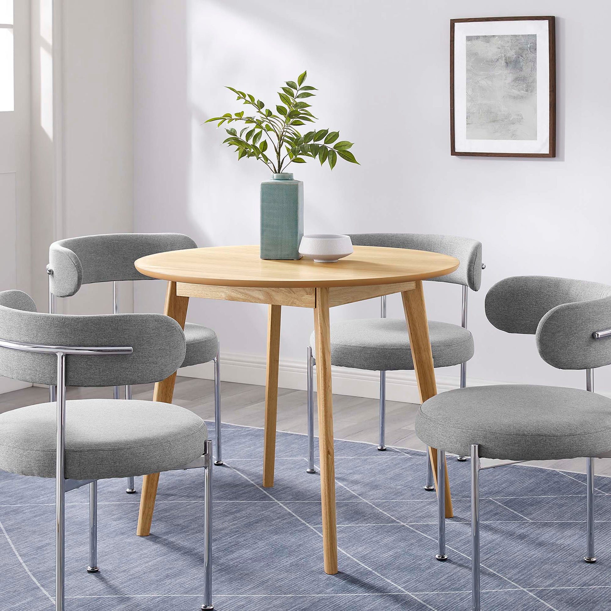 Round oak dining table with tapered legs, surrounded by light gray upholstered chairs; plant vase centerpiece on blue rug.