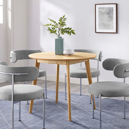 Round oak dining table with tapered legs, surrounded by light gray upholstered chairs; plant vase centerpiece on blue rug.