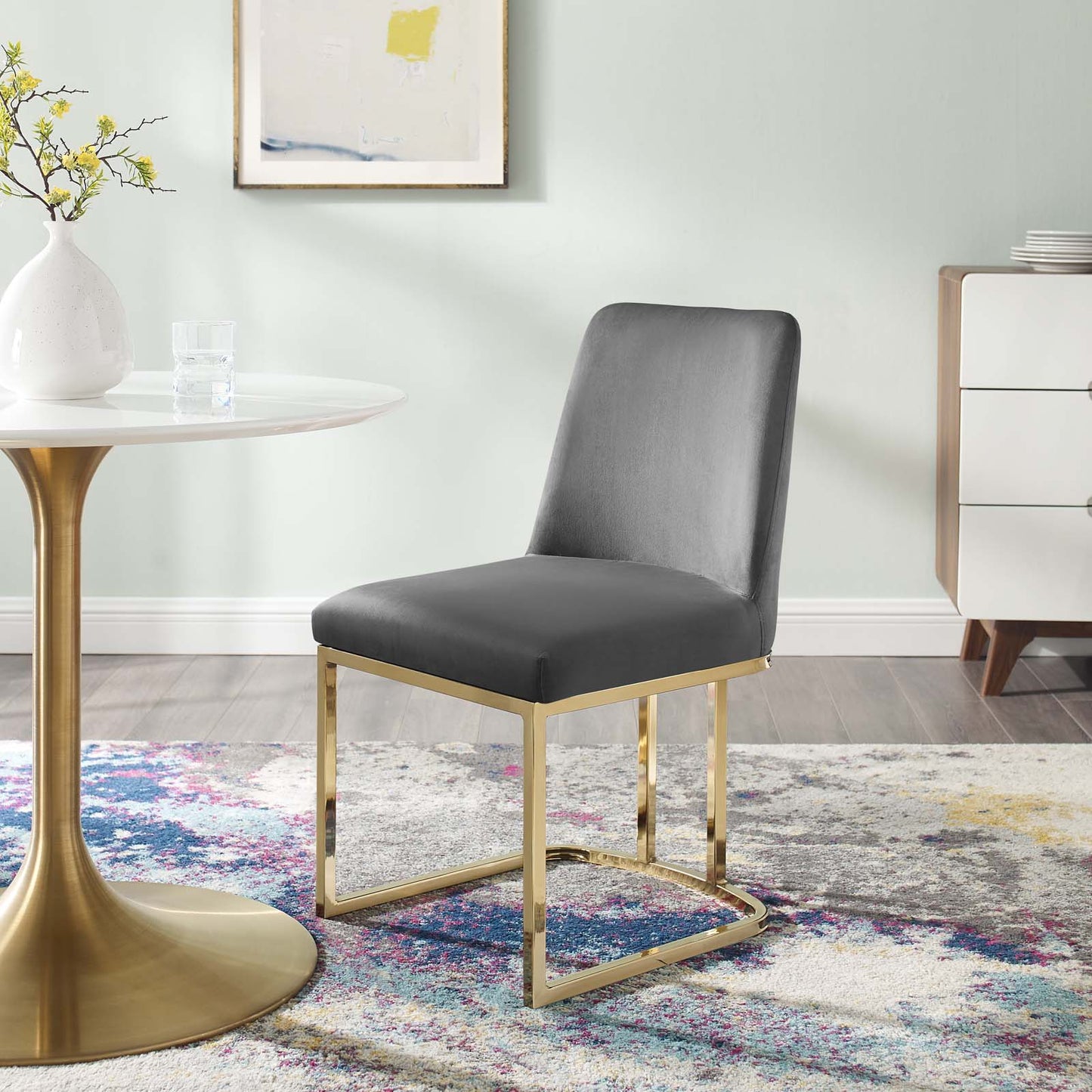 Grey upholstered dining chair with a gold-tone sled-base beside a round glass table in a modern pastel dining room.
