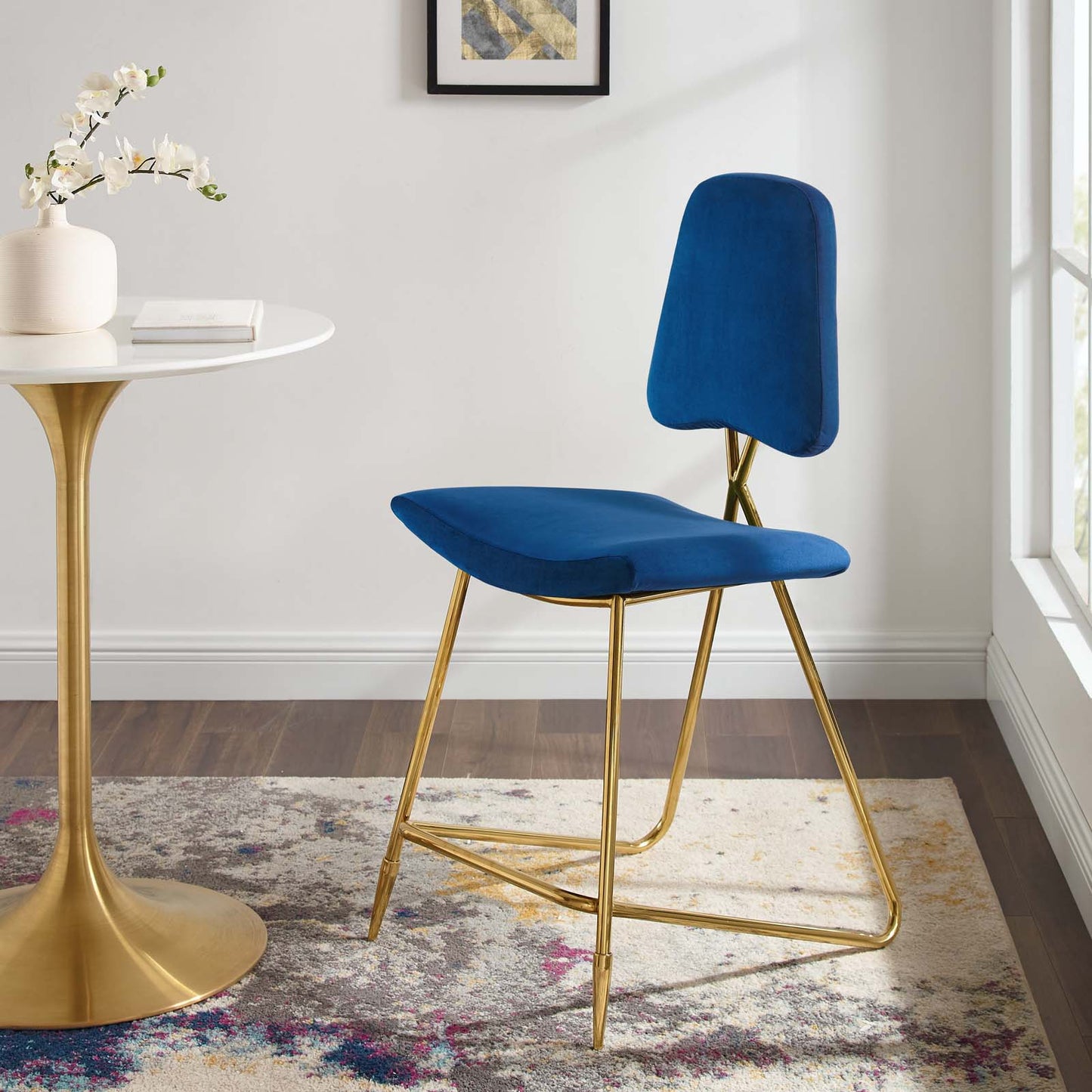 Navy blue velvet counter stool with slim gold metal legs beside a white round table and vase in a bright modern room.