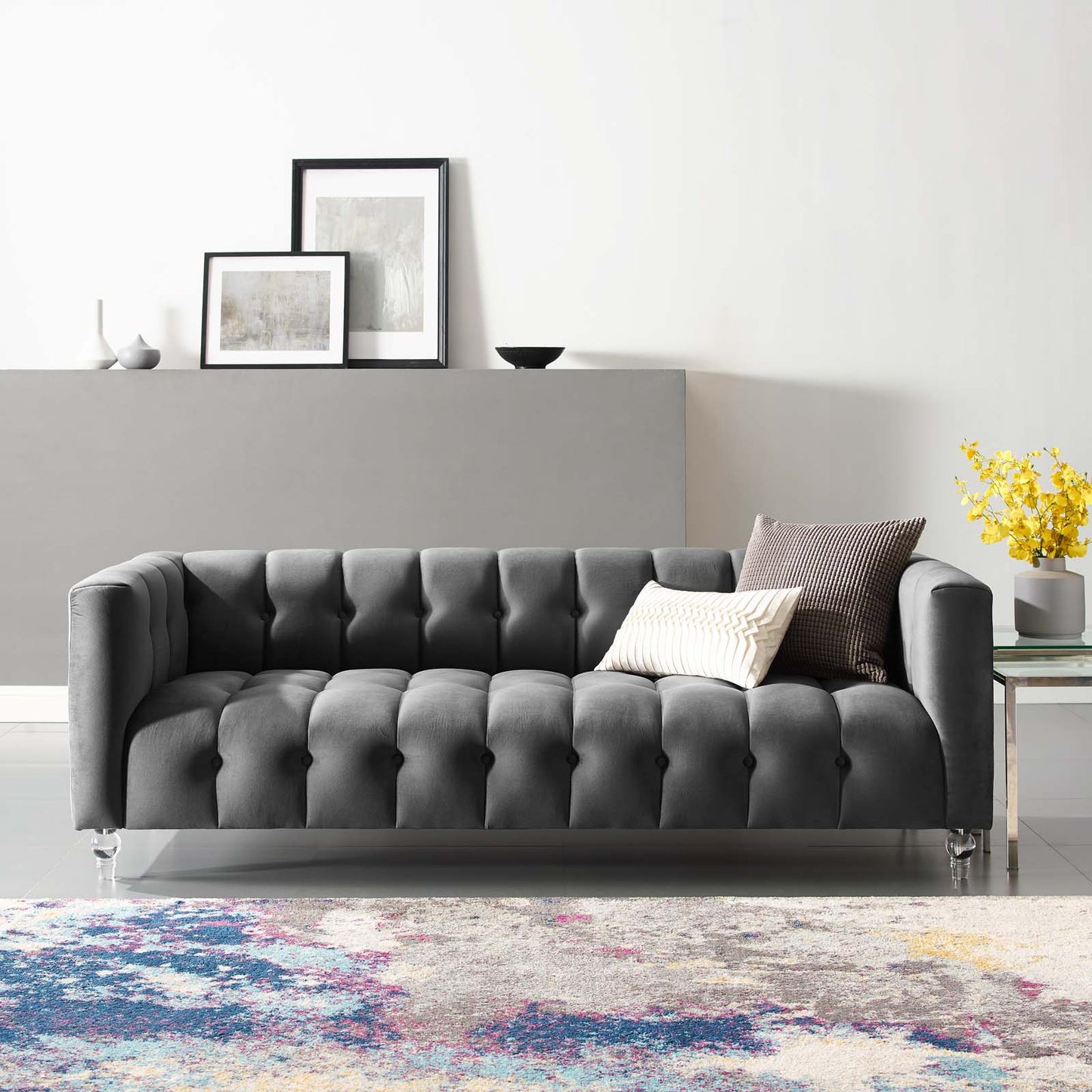Grey tufted sofa with deep button tufting and slim legs, two cushions, in a bright living room with a colorful rug.