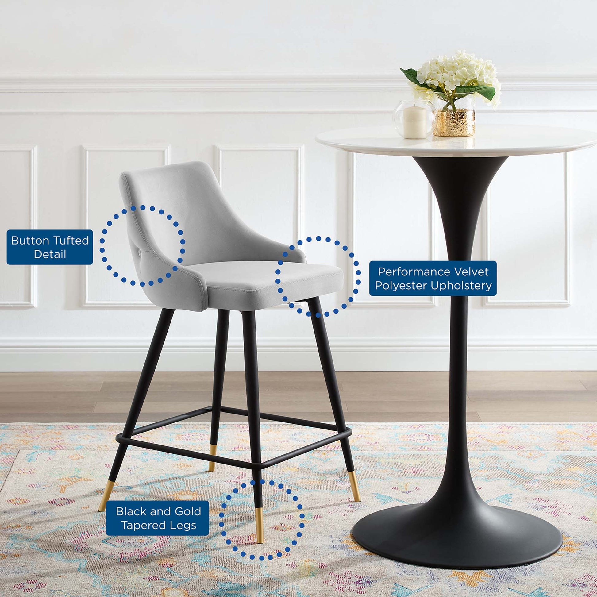Light gray button-tufted velvet counter stool with black tapered legs and gold tips beside a white round pedestal table.