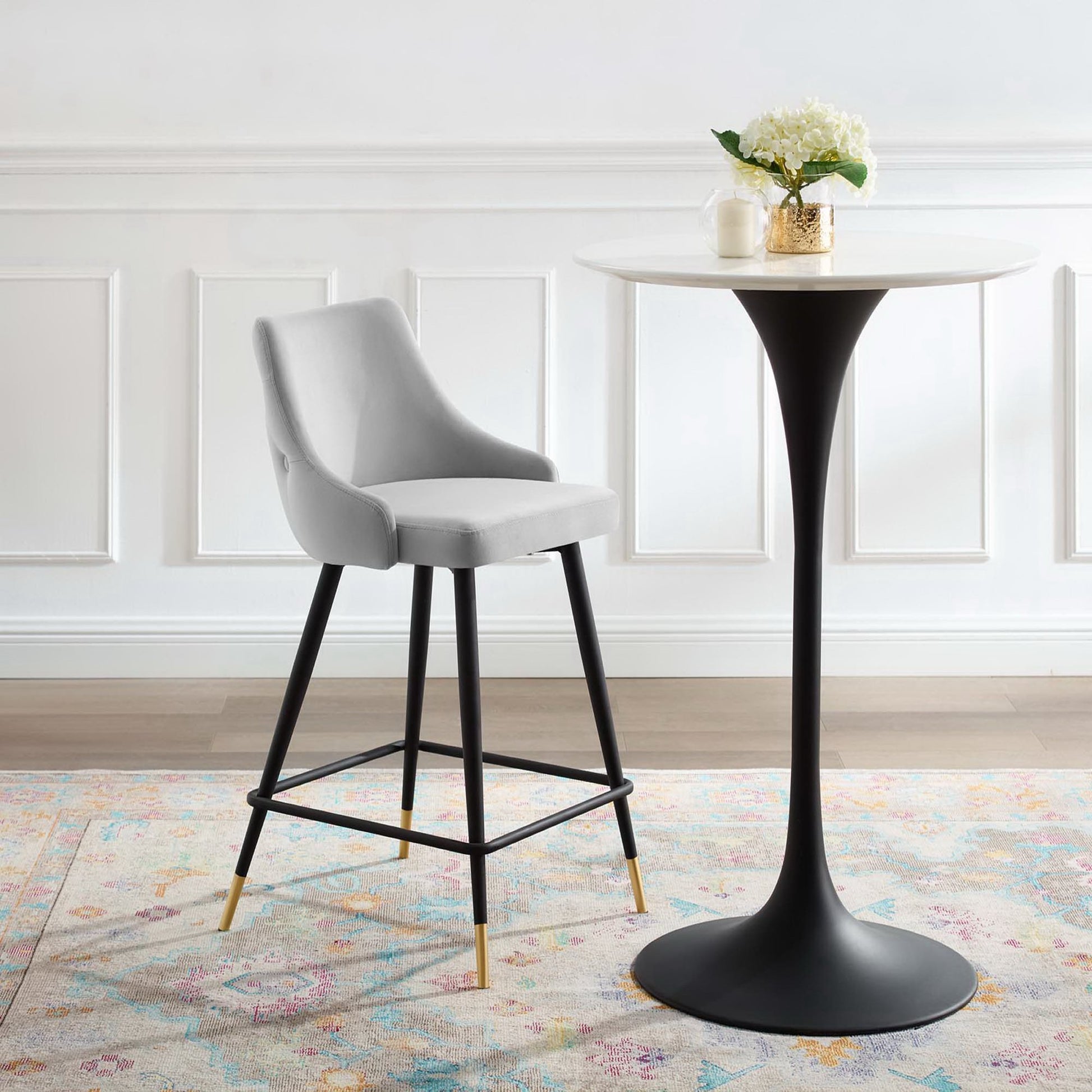 Gray upholstered counter stool with black legs and brass tips beside a white round pedestal table in a bright modern dining nook.