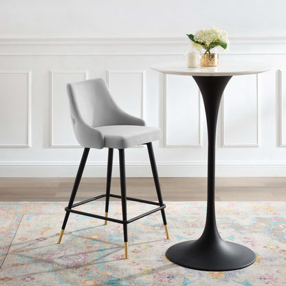 Gray upholstered counter stool with black legs and brass tips beside a white round pedestal table in a bright modern dining nook.