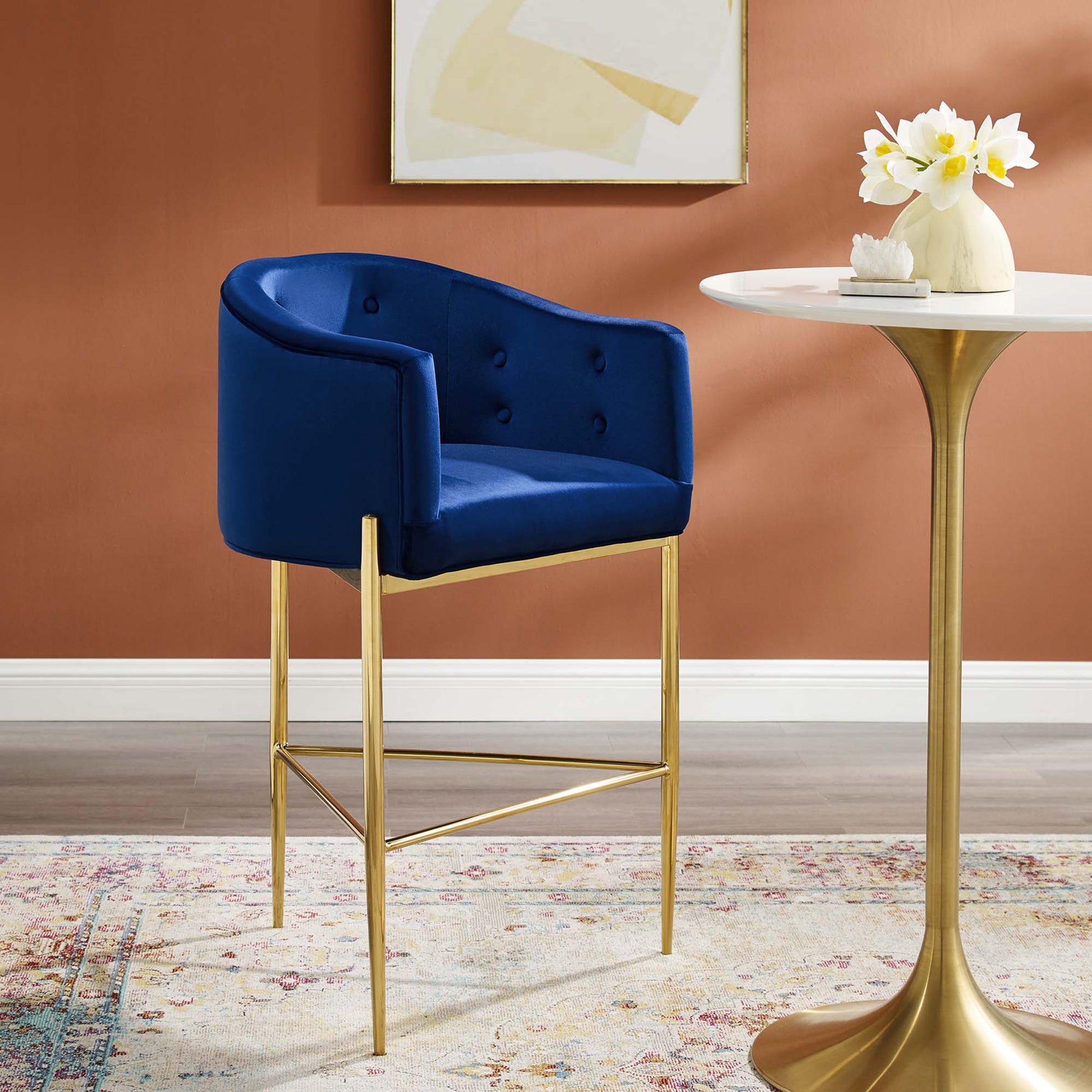 Navy tufted velvet bar stool with gold metal legs, shown beside a round marble-top table with flowers.