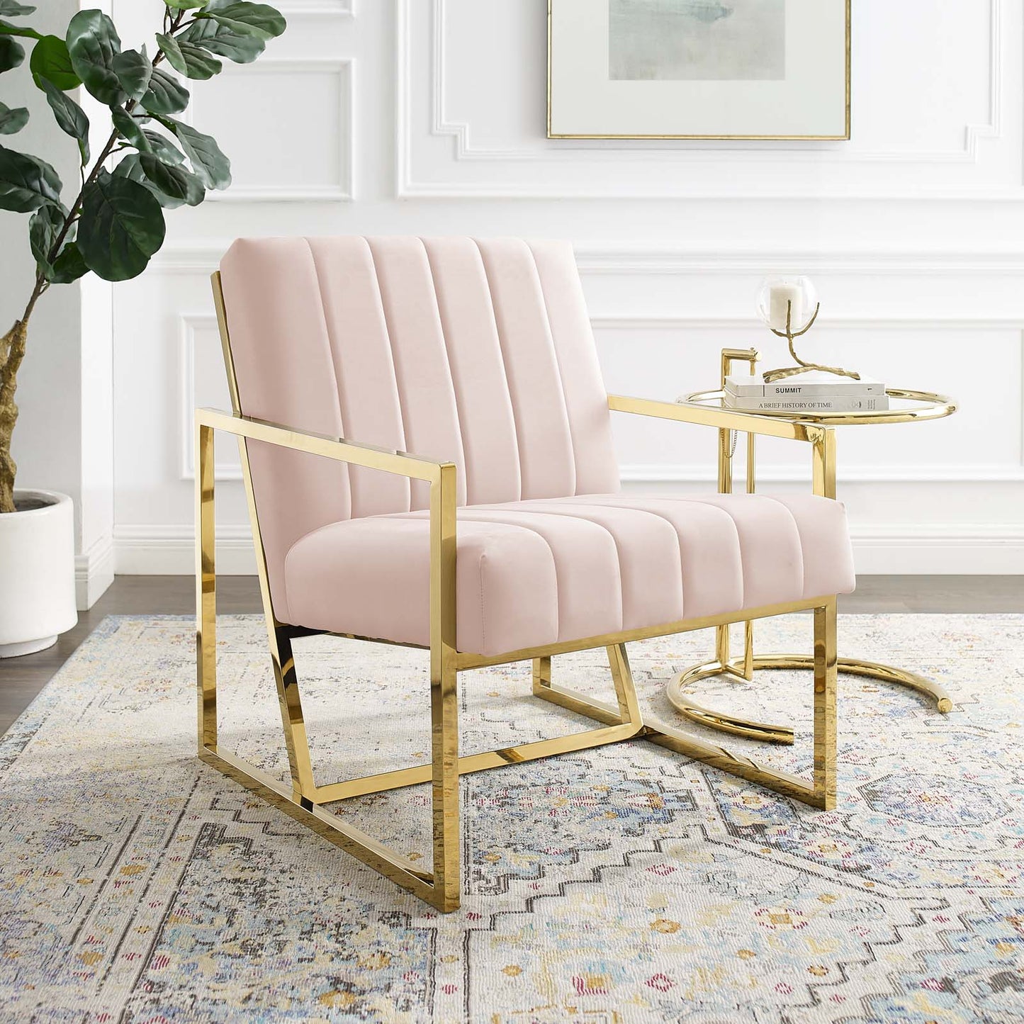 Pink velvet armchair with vertical channel tufting and a gold metal frame beside a round gold side table.