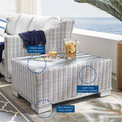 Light gray synthetic rattan outdoor coffee table on a seaside patio; blue blanket and glass pitcher.
