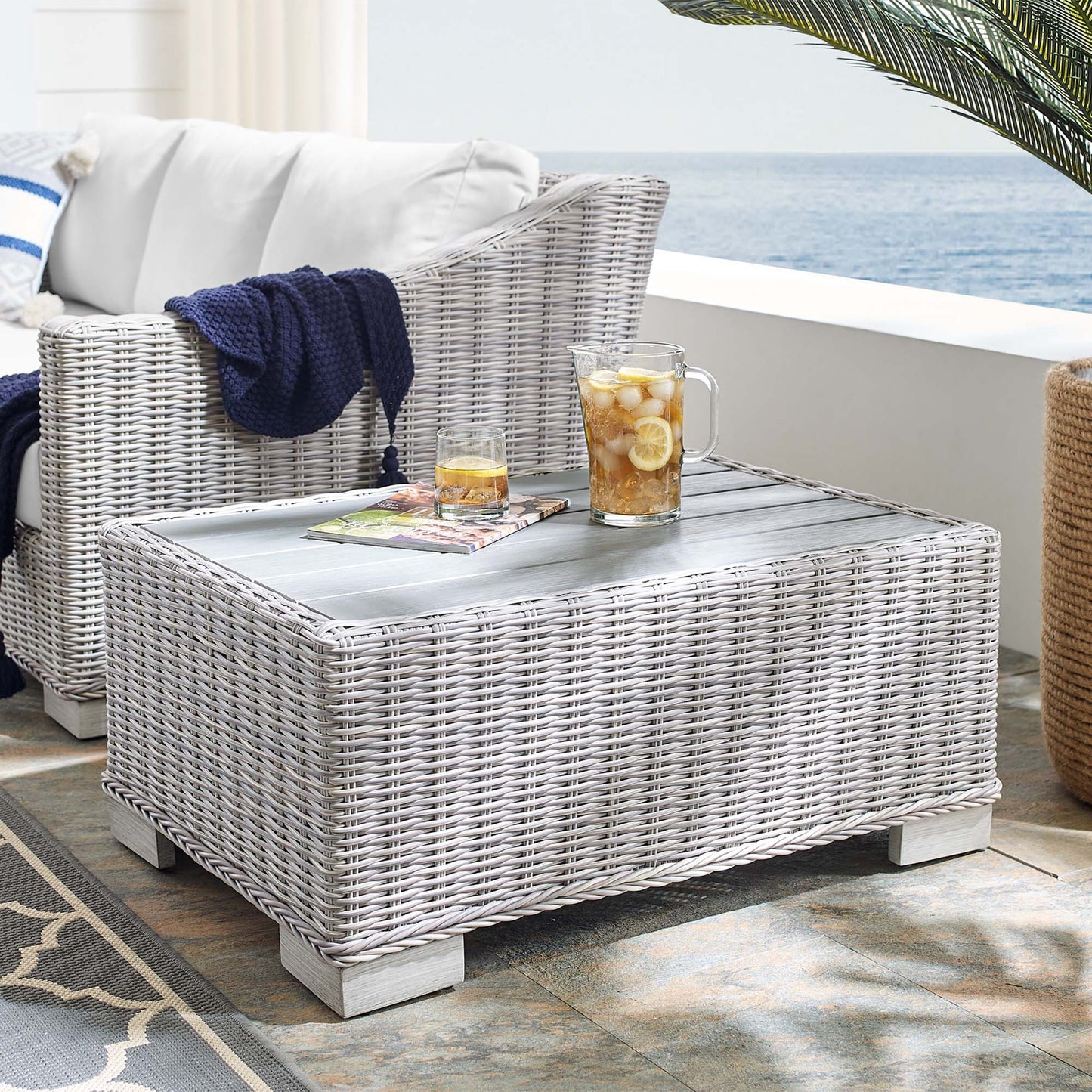 Rectangular light gray wicker outdoor coffee table with lemonade pitcher, glass, and magazine on a sea-view patio.