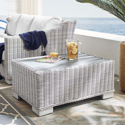 Rectangular light gray wicker outdoor coffee table with lemonade pitcher, glass, and magazine on a sea-view patio.