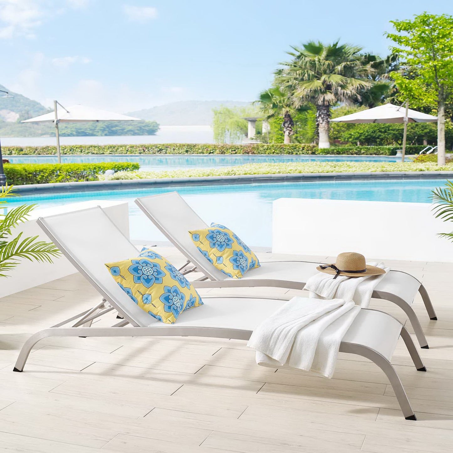 Two white curved poolside chaise loungers with blue floral cushions, white towel, straw hat, by a sunny resort pool.