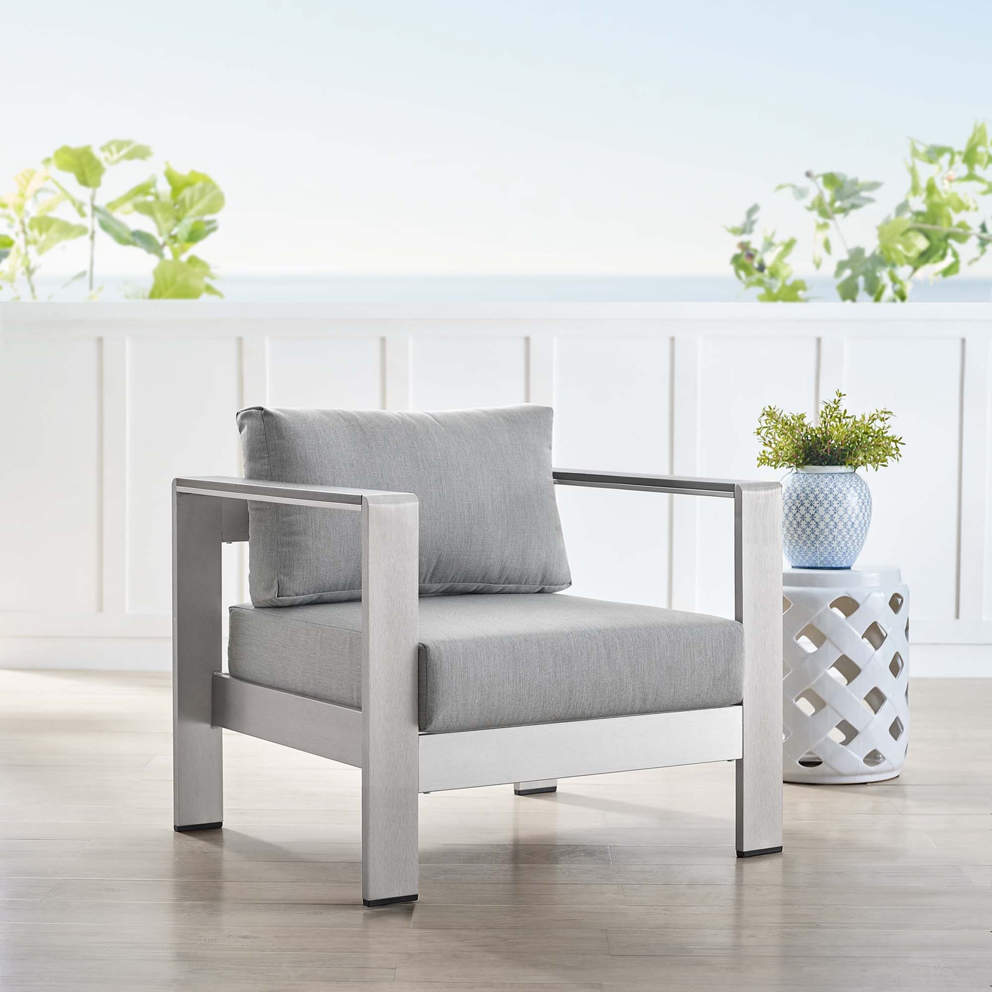 Modern outdoor armchair with a silver-gray aluminum frame and light gray cushions on a sunlit deck, with a lattice side table and a potted plant.