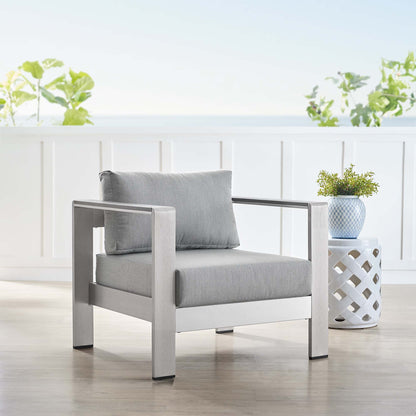 Modern outdoor armchair with a silver-gray aluminum frame and light gray cushions on a sunlit deck, with a lattice side table and a potted plant.