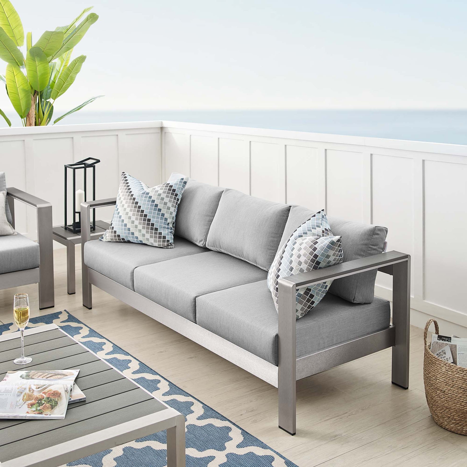 Slate gray outdoor sofa with light gray cushions and geometric-pattern pillows on a modern metal-frame patio set by the sea.
