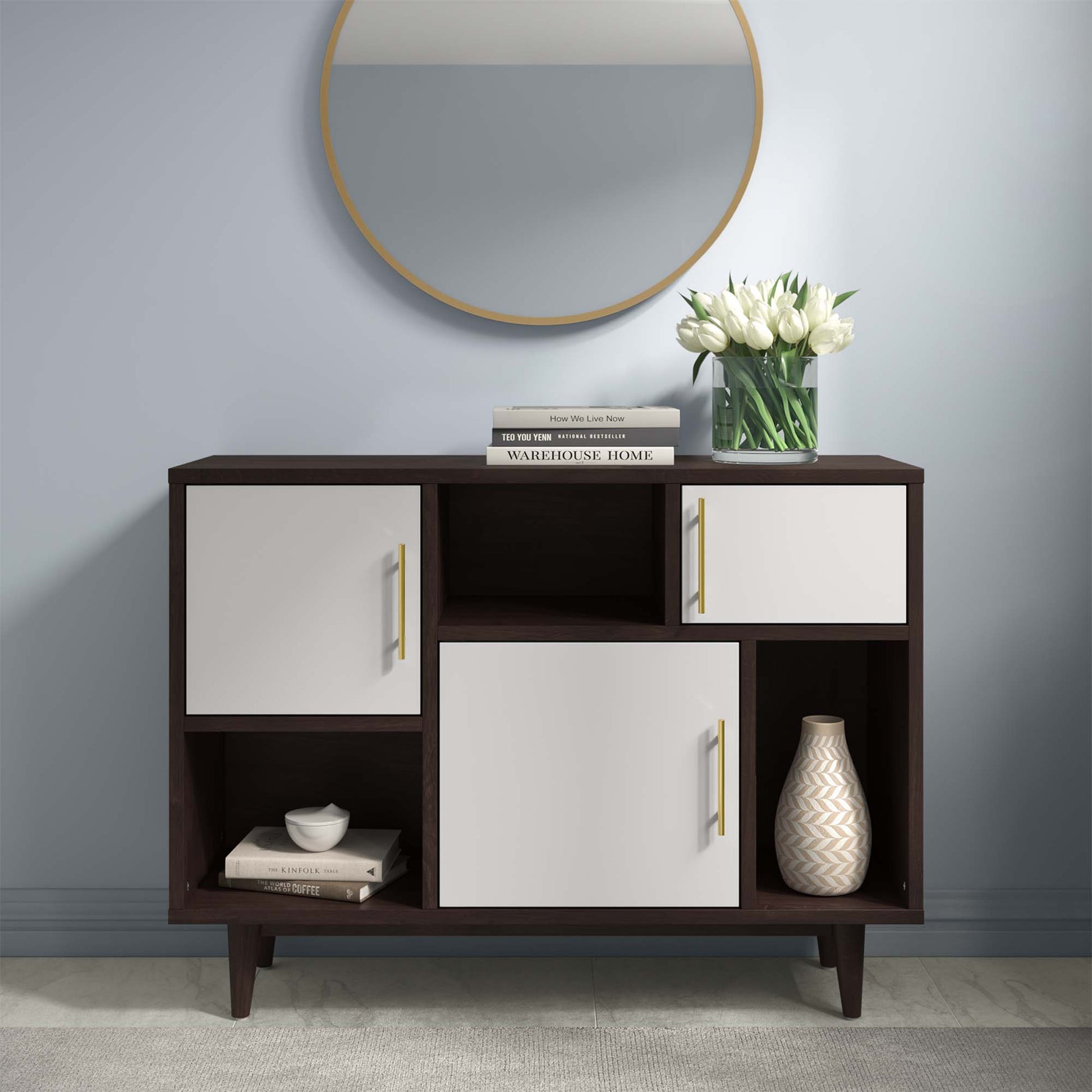 Display stand in cappuccino white finish with dark wood tones, white panel doors with gold pulls, open shelves; gold-framed mirror, vase and books on top.