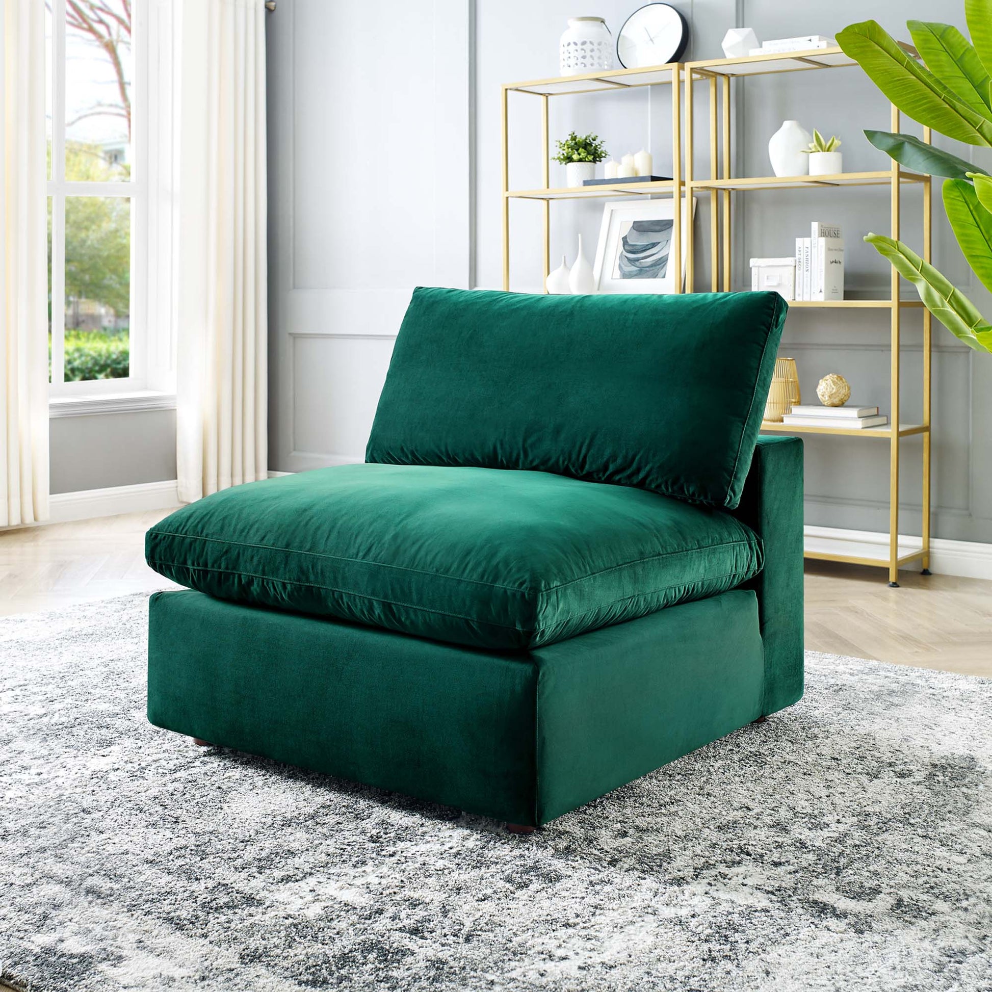 Emerald green velvet armless chair with thick cushions in a modern living room with gold shelving and a large plant.
