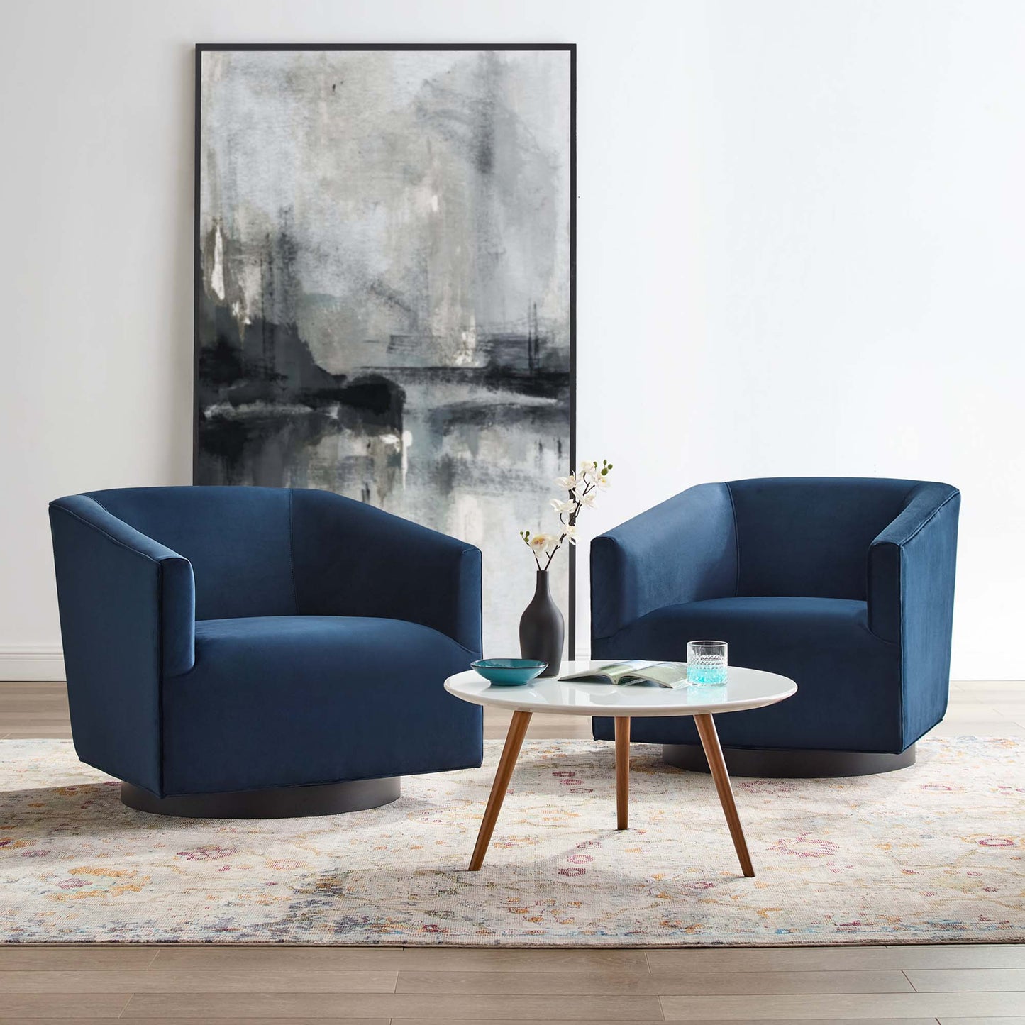 Two navy blue velvet swivel chairs with wooden bases surround a white round coffee table; abstract art, vase, patterned rug.