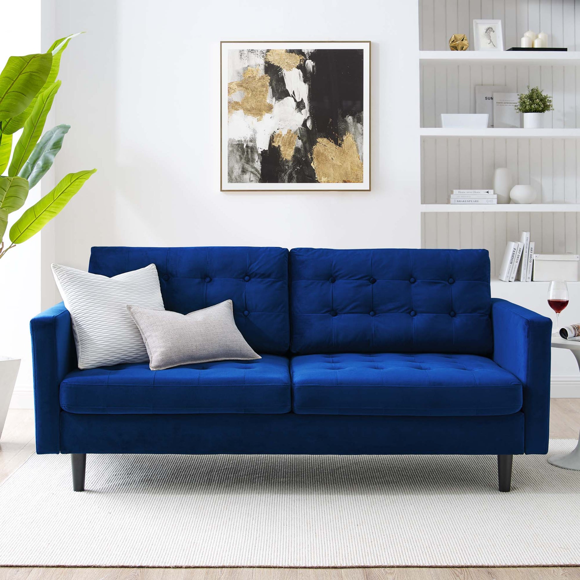Navy blue velvet tufted sofa with two light cushions on slim black legs in a bright modern living room with abstract wall art.