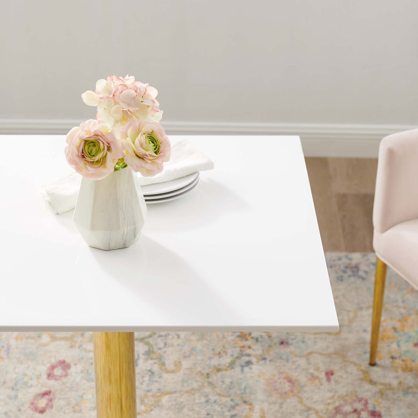 White square dining table with natural wood legs, geometric vase with pink & cream flowers, plates and napkin, pink chair nearby.