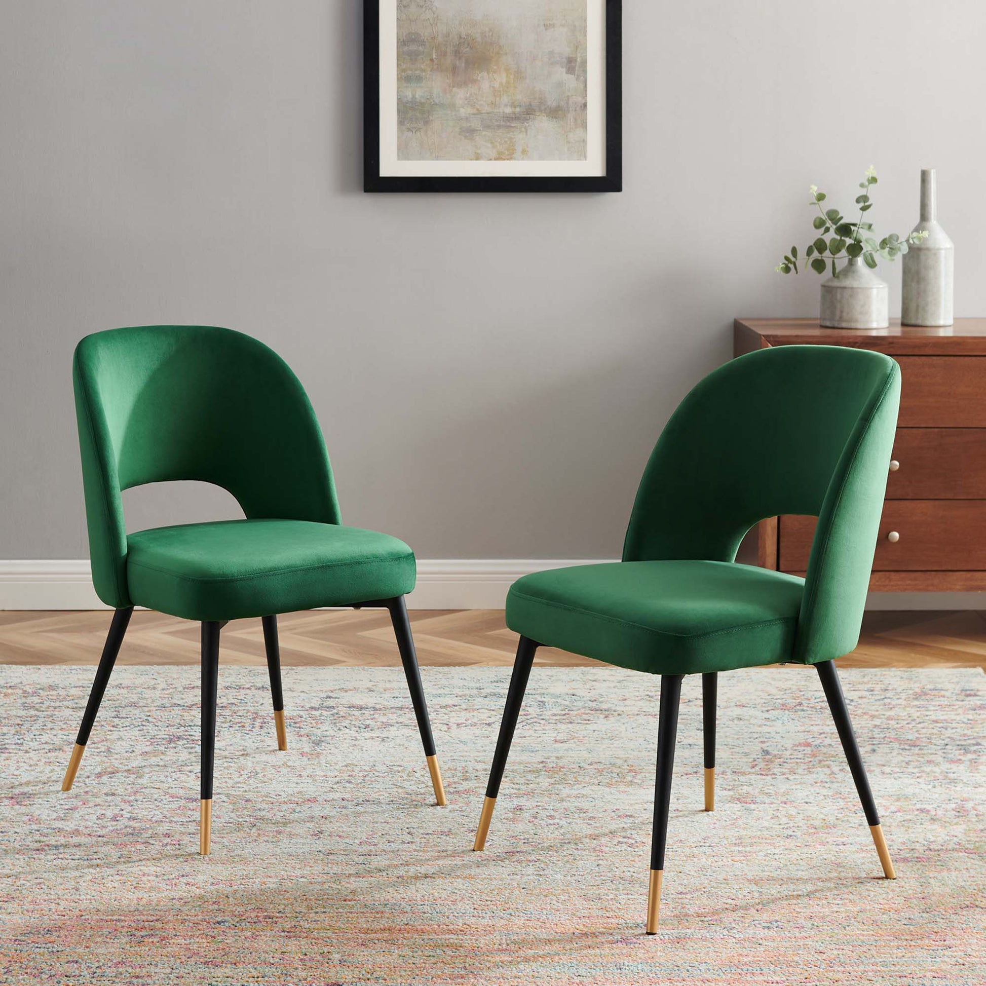 Two emerald green velvet dining side chairs with curved backs and black metal legs with gold tips, chic mid-century style.