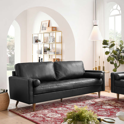 Black leather sofa with wooden legs and cylindrical arm bolsters; modern living room with a gold metal bookshelf and red rug.