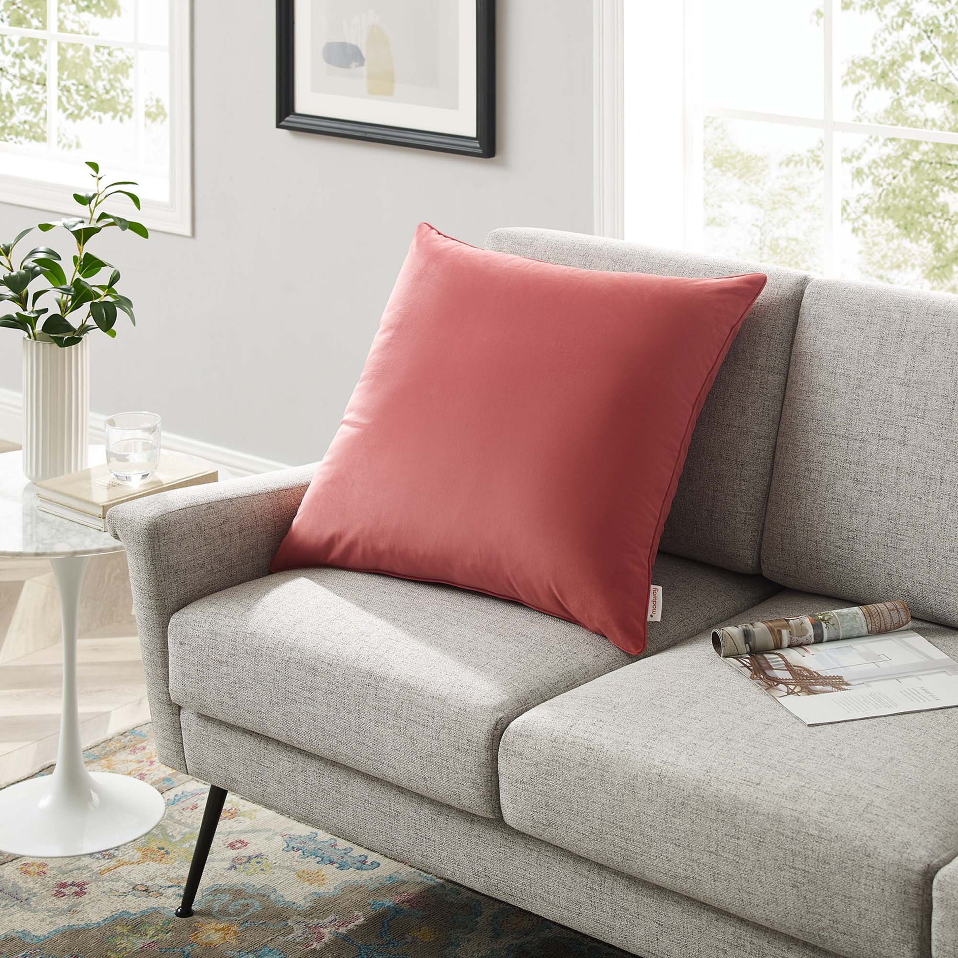 Coral pink square decorative pillow on light gray textured sofa in a bright modern living room with a plant.