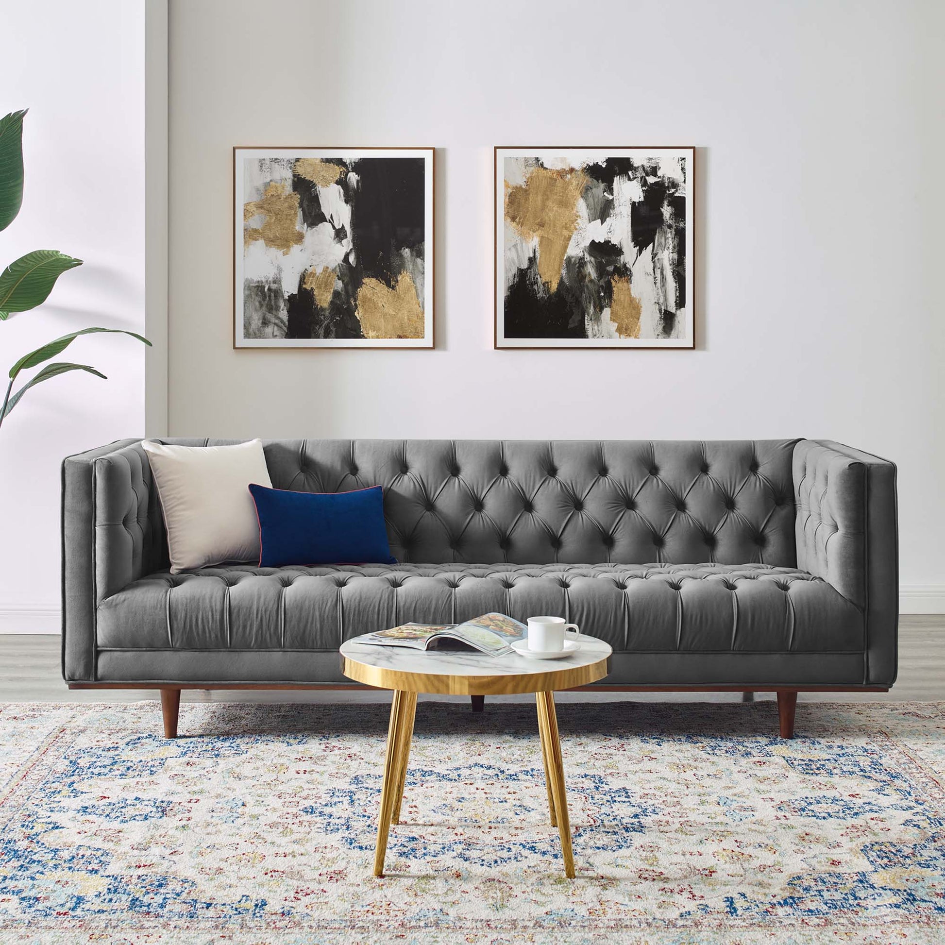 Gray tufted sofa with wooden legs, beige and blue cushions; abstract gold-black art above; round wooden coffee table on rug.
