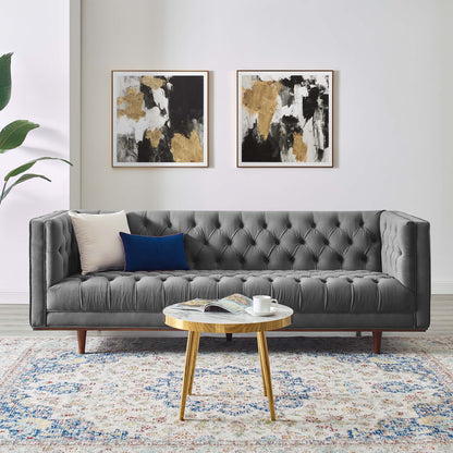Gray tufted sofa with wooden legs, beige and blue cushions; abstract gold-black art above; round wooden coffee table on rug.