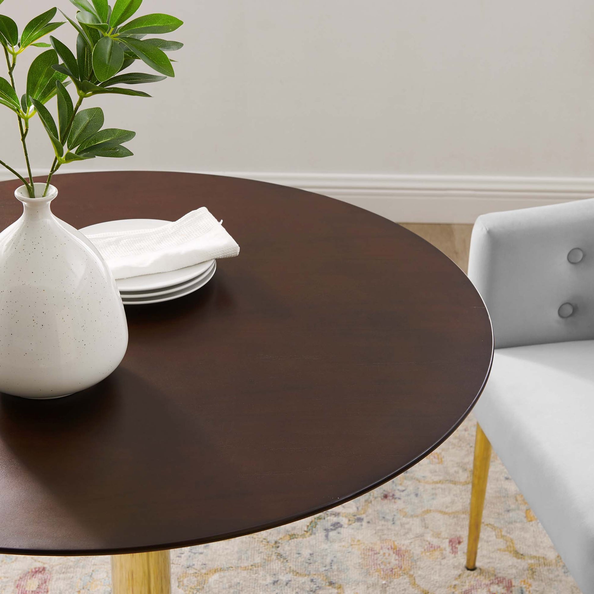 Round dark wood dining table with white speckled ceramic vase, green foliage, plates and napkin; gray upholstered chair.