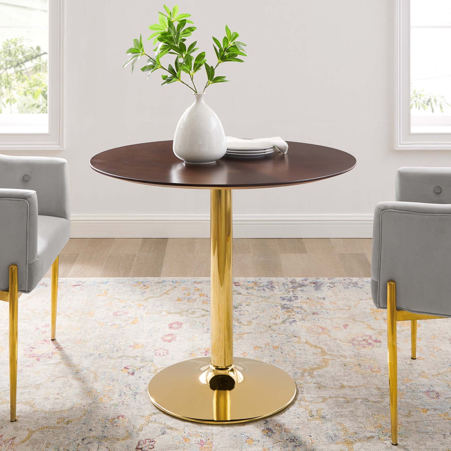 Round dark wood table with gold pedestal base, white vase with green plant, stack of plates; gray chairs with gold legs.