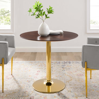 Round dark wood table with gold pedestal base, white vase with green plant, stack of plates; gray chairs with gold legs.