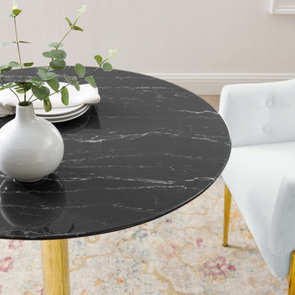 Round black marble table with white veining on golden wooden legs, vase with greenery and stacked plates beside a chair.