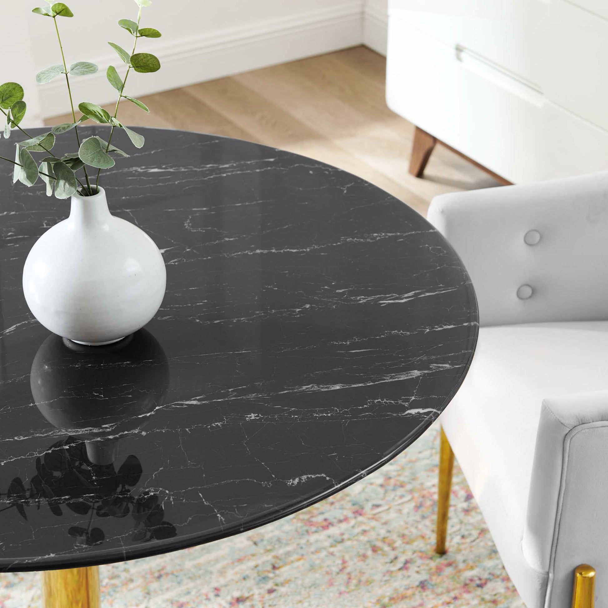 Round black marble tabletop with white veining, white ceramic vase with greenery, white chair with gold legs in living room.