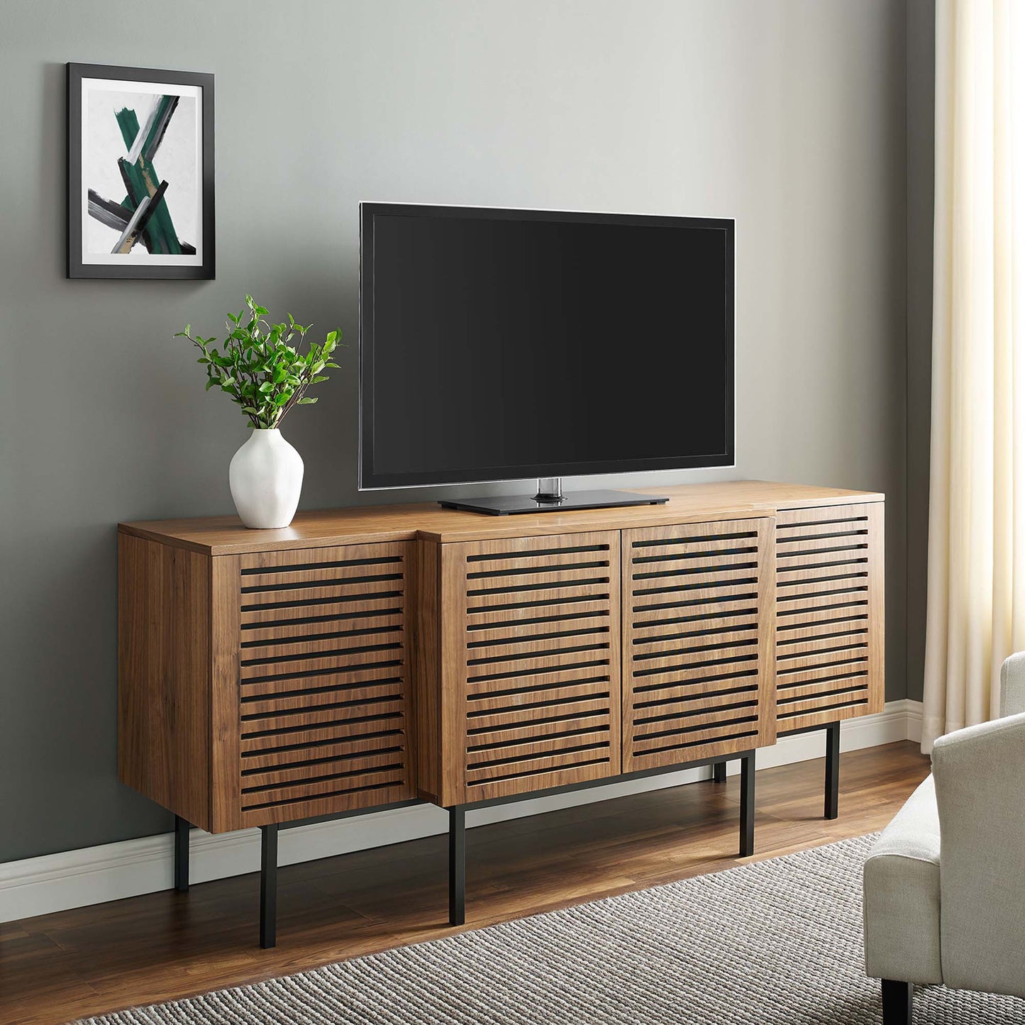 Walnut sideboard with slatted cabinet doors, slim black legs, TV on top, white vase with greenery and framed art