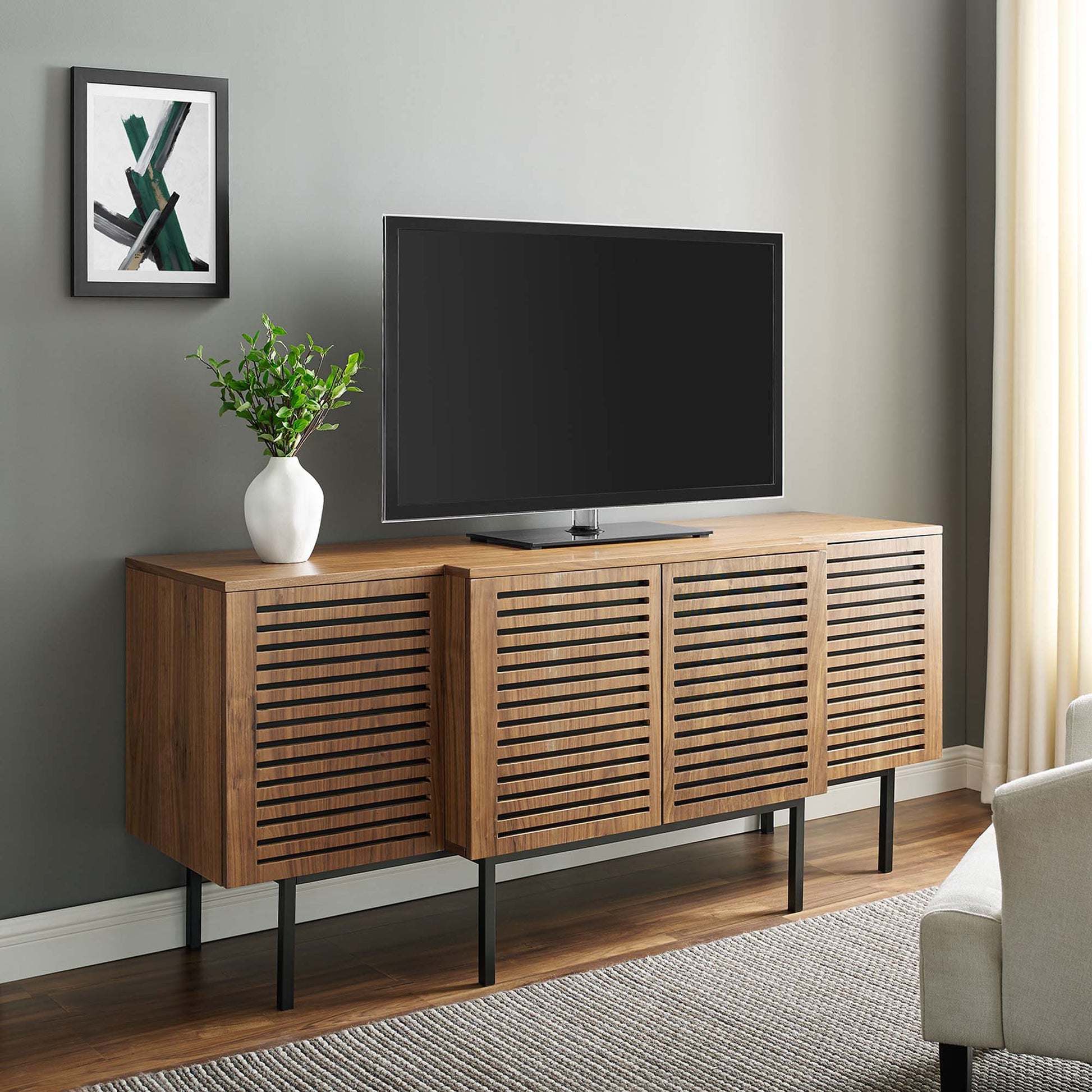 Walnut sideboard with slatted cabinet doors, slim black legs, TV on top, white vase with greenery and framed art