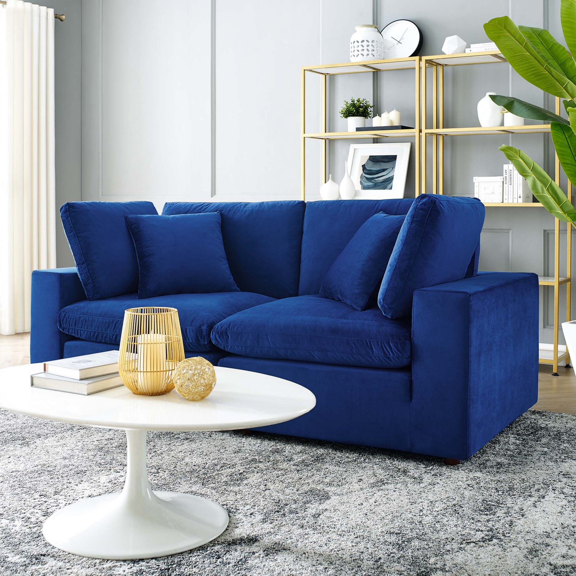 Navy velvet loveseat with plush cushions in a modern living room, white round coffee table and gold shelving.