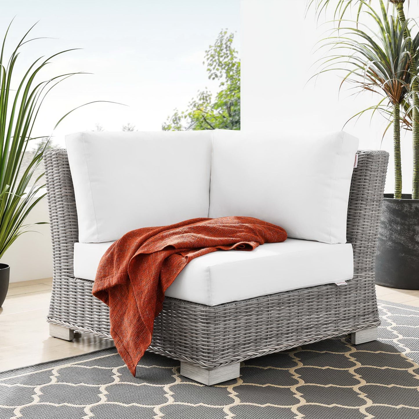 Gray synthetic wicker outdoor corner chair with white cushions and an orange throw on a patterned patio rug.