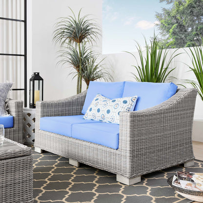 Outdoor grey wicker loveseat with light blue cushions and a patterned lumbar pillow, on a lattice rug among potted plants.