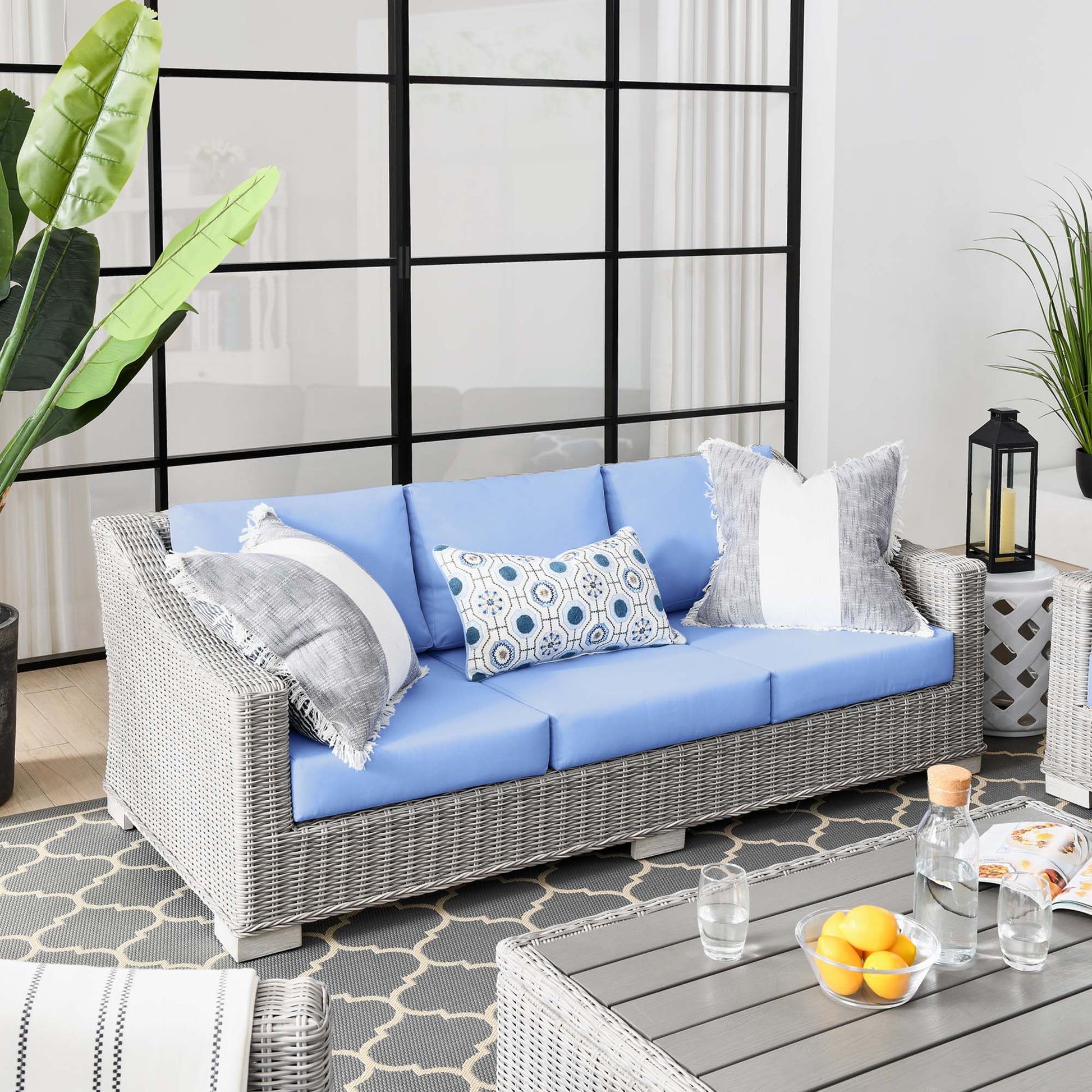 Light gray wicker outdoor sofa with light blue cushions, decorative pillows, throws, on a patterned rug in a sunlit patio.