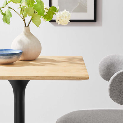 Natural wood table on black pedestal leg with blue ceramic bowl, beige vase with green leaves and white flower, gray chair.
