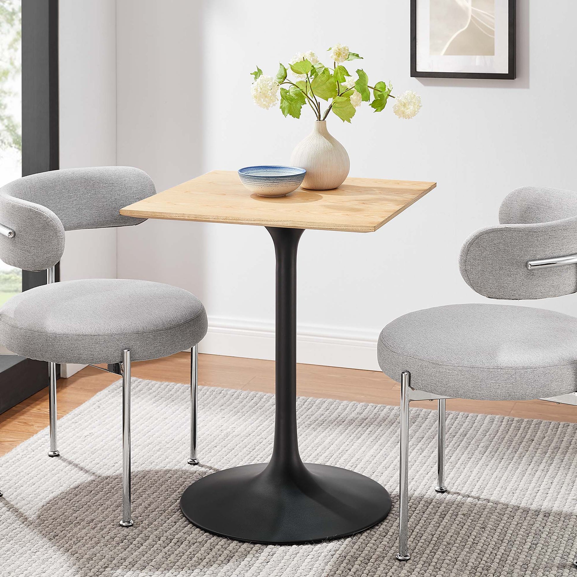 Natural wood square-top dining table on a black tulip pedestal, with two grey upholstered chairs; vase of white flowers and blue bowl.