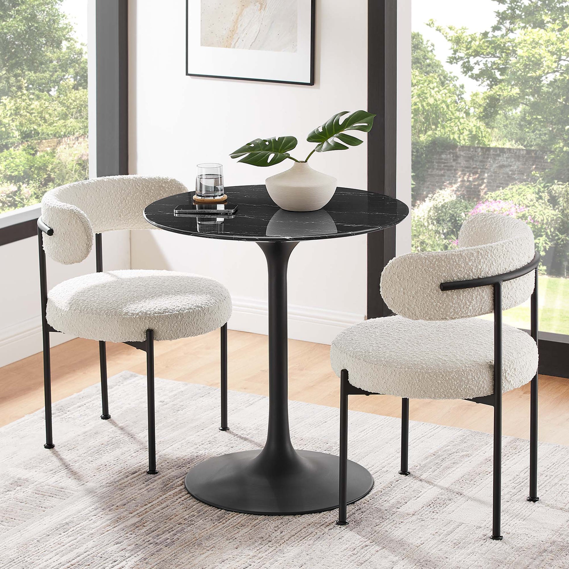 Round black faux-marble dining table with a pedestal base, accompanied by two white boucle chairs on black metal legs in a sunlit dining nook.