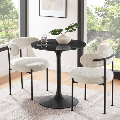Round black faux-marble dining table with a pedestal base, accompanied by two white boucle chairs on black metal legs in a sunlit dining nook.