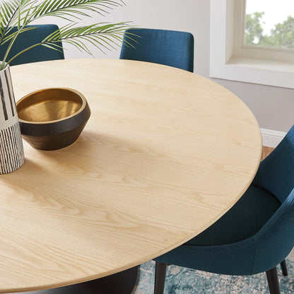 Round natural wood dining table with blue upholstered chairs; black and gold bowl, striped vase, and green plant.