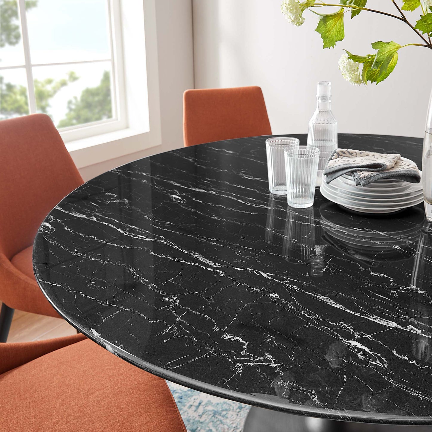 Round black faux-marble dining table with white veining, orange chairs, glassware & stacked plates in a bright dining area.