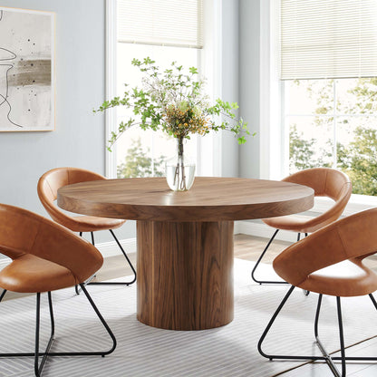 Round walnut dining table with a thick pedestal base, surrounded by caramel leather chairs and a greenery vase centerpiece.