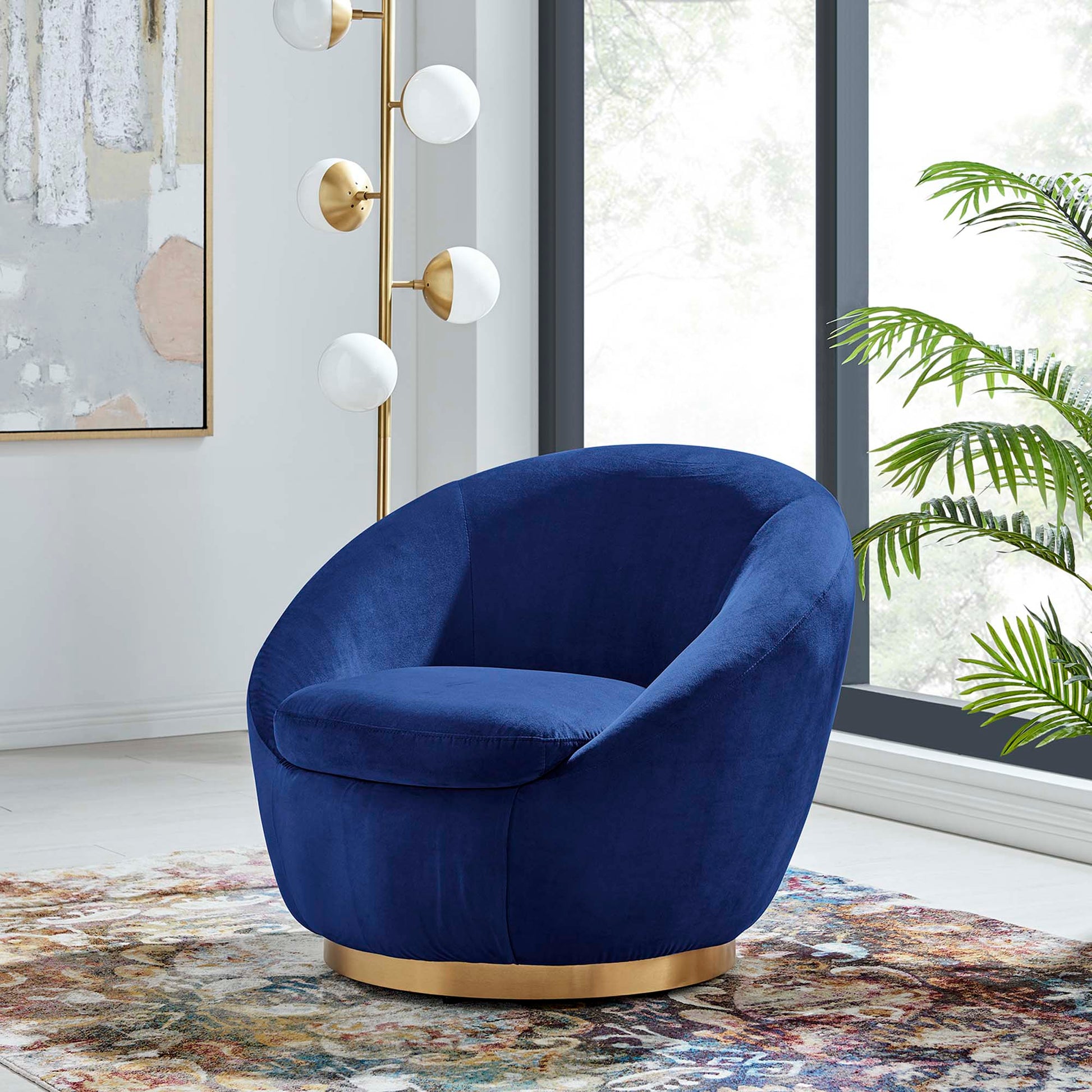 Navy velvet curved swivel chair with a gold base in a bright living room; globe floor lamp, plant & colorful rug.